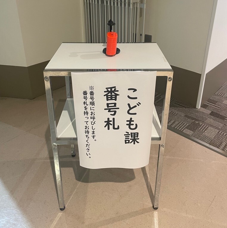 2階エレベーター前にこども課番号札と書かれた台と台の上に赤い番号札が順番に重なっています。